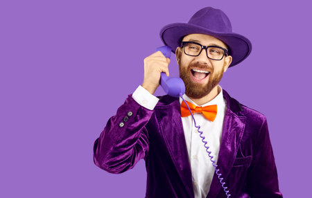 Smiling showman isolated on purple studio background talk on corded phone with client. Happy man in disco suit and hat have pleasant landline telephone call. Advertising, copy space.の写真素材