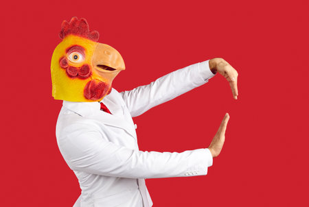 Man wearing strange chicken mask having fun at crazy animal party. Side profile view shot of guy with funny rooster head dancing isolated on solid red colour backgroundの写真素材