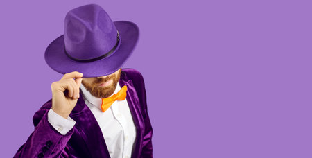 Handsome man with ginger beard, wearing hat, suit and orange bowtie, raises his fedora hat a bit as he bows his head standing on blank solid purple banner background with copy space on right sideの写真素材