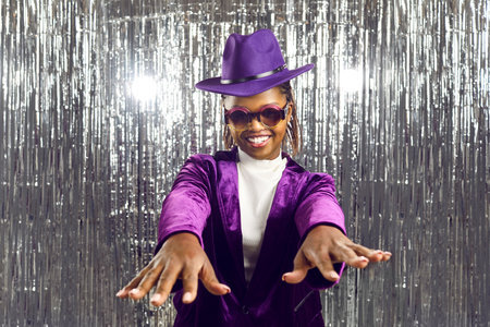 Happy black woman having fun at a party. Portrait of a beautiful young African American girl in a purple hat, velvet suit and disco glasses dancing on a sparkling silver foil fringe backgroundの写真素材