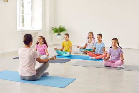 Portrait of a little kids girls sitting on the floor with their teacher and practicing yoga sitting in gym in Lotus pose with closed eyes. Children sport and active lifestyle concept.の写真素材