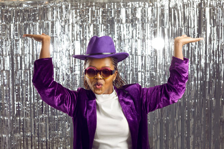 Young positive dark-skinned woman in corduroy bright costume performs funny dance moves on silver shiny background. Carefree woman with funny expression dances with raised hands. Party concept. Bannerの写真素材