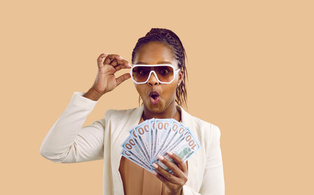 Happy rich wealthy African American woman in cool sunglasses on beige studio background holding fan of paper money. Joyful excited lady earns a lot and looks at first salary in hands with shockedの写真素材
