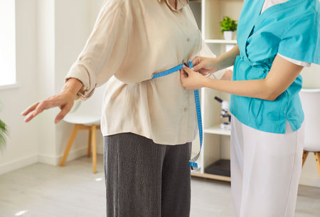 Senior patients waist measurement by nurse holding measuring tape on medical checkup in hospital. Older woman standing with arms out to measure waist on health screening, obesity preventionの写真素材