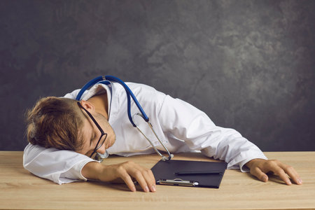 Studio portrait of tired and exhausted young male medical worker sleeping sitting at table. Overworked medic on gray background. Concept of stress and fatigue of medical staff.の写真素材