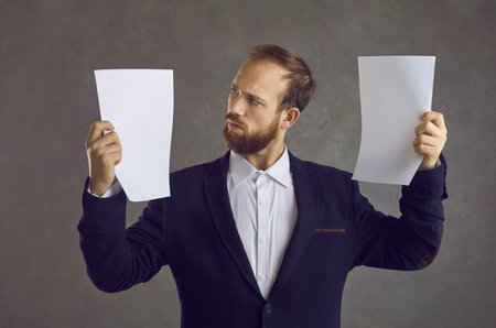 Studio portrait of adult caucasian businessman comparing two document holding in hands. Professional accountant, analyst or adviser looking at blank paper sheets standing over grey. Business conceptの写真素材