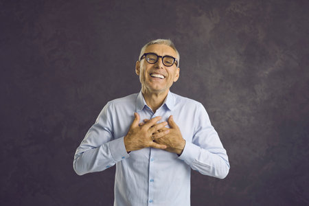 Thank you from the bottom of my heart. Happy smiling senior Caucasian man holding hands on chest. Cheerful old businessman, pensioner or grandfather expressing endless gratitude for present or giftの写真素材