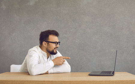 Side view of a happy young man in glasses pointing at a PC laptop. Handsome guy sitting at a desk, smiling, looking and pointing at the computer screenの写真素材