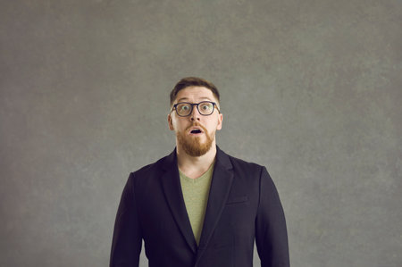 Studio portrait of scared or shocked young guy. Man in suit jacket and glasses looking at camera with expression of surprise, shock or fear on his faceの写真素材