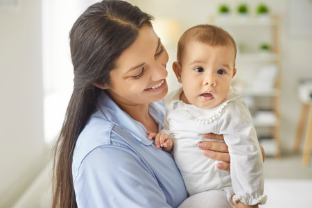 A smiling mother holds her curious baby toward the camera in a bright bedroom, making a warm...