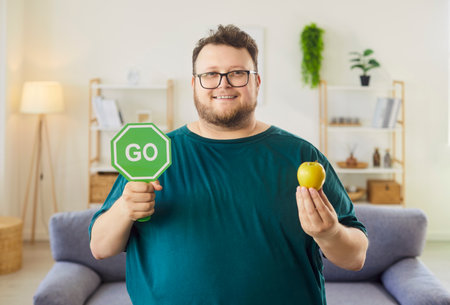 Man with GO sign presents healthy apple. In a bright living room, the smiling male promotes smart nutrition and steady diet, holding a green octagon and fruit. Concept: go for healthy choices.の写真素材