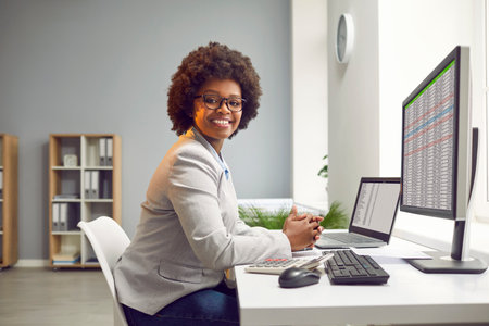 Side view portrait of a happy smiling african american business woman accountant sitting at the desk and working on a laptop with tables and charts at office or at home and looking at the camera.の写真素材