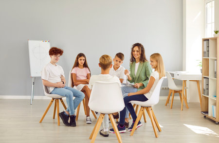 Friendly young school psychologist woman having conversation with a group of cute children sitting in a circle in classroom. Psychological support for kids and mental health concept.の写真素材
