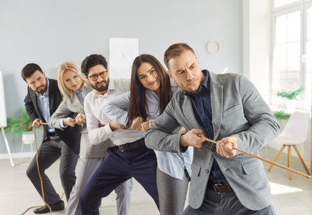 Business team in formal attire playing tug of war with rope in office. Competitive colleagues having fun together and engaging in team building activity while strengthening their collaboration.の写真素材