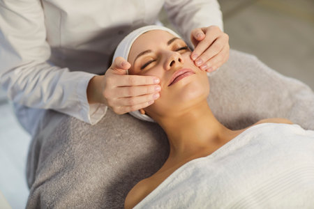 Top view of young woman enjoying soothing facial massage during skincare procedure. Professional cosmetologist gently massaging face of female client, providing relaxing beauty treatment in spa salon.の写真素材