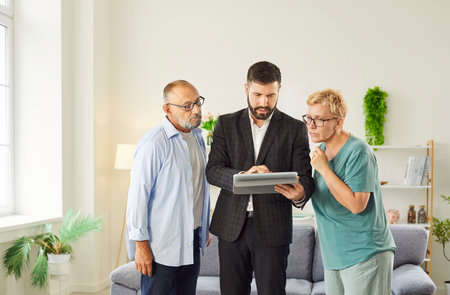Realtor agent reviews contract and home purchase details with a mature senior couple reviewing financing and showing documents on a tablet in living room. Male advisor consulting elderly people.の写真素材