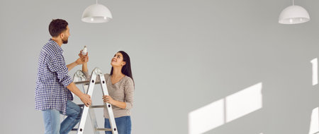 Young man standing on the ladder changing light bulb in the room with his smiling wife. Happy couple installing the chandelier in their new empty apartment. Banner.の写真素材