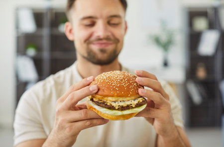 Close-up of young 30s smiling man holding juicy, freshly prepared, appetizing, homemade or delivered hamburger alone at home. Concept of meal enjoyment, junk food habits lifestyleの写真素材