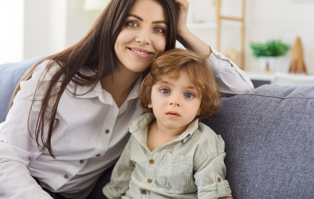 Happy young woman cuddling small cute kid son portrait, having fun together on couch in living room, child boy nanny babysitter, laughing at home, looking at camera, posing for family photoの写真素材