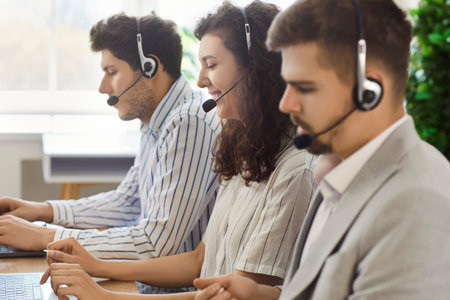Three call center employees wearing headsets sitting at desk, using laptops and providing voice support to clients. Customer service operators handling service requests and providing customer care.の写真素材