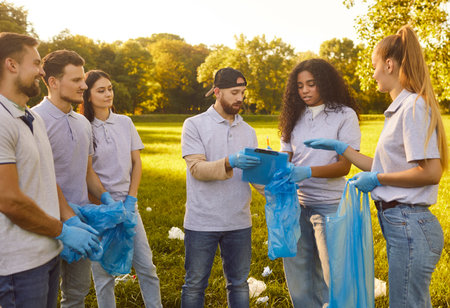 Group of young people volunteers in gloves holding trash bags working in summer park collecting garbage outdoors. Men and women cleaning environmental pollution in nature. Ecology concept.の写真素材