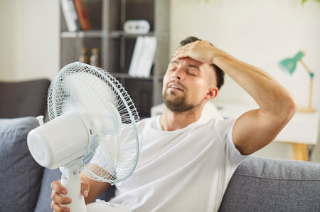 Tired man with electric fan in hot home. Seated on sofa, he seeks cooling airflow suffering from summer heat, wiping sweat and trying to relax. Unhappy frustrated male enjoying cool air.の写真素材