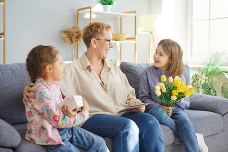Happy grandchildren giving greeting card, present box and flowers bouquet to senior elderly...