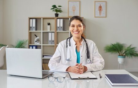 Portrait of cheerful young friendly female doctor in white medical uniform sitting at the desk on workplace with laptop. Happy woman physician wearing stethoscope looking at camera and smiling.の写真素材