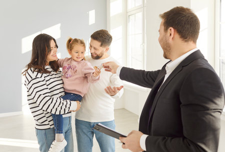 Portrait of man realtor giving house keys to a happy smiling young family of mother, father and child girl standing in empty new apartment. Real estate, moving and buying property concept.の写真素材