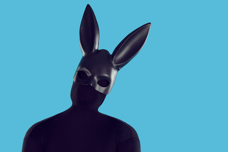 Portrait of man disguised in rabbit costume in fashion studio. Guy in faceless skintight monochromatic black bodysuit and long eared bunny mask tilts head, standing like silhouette on blue backgroundの写真素材