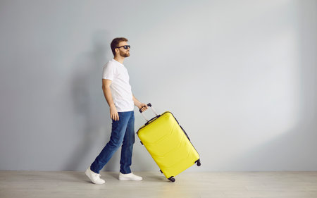 Male traveller with luggage. Side view funny man in white T shirt, blue jeans and sunglasses walking with yellow suitcase along light grey wall background. Travelling, holiday, vacation, trip conceptの写真素材