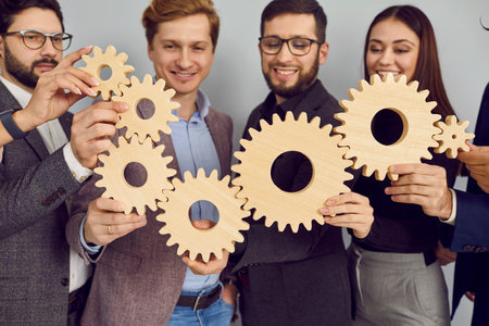 Business team holding gears to show concept of teamwork and integration. Group of happy young people standing in office, making chain of wooden cog wheels and smiling. Working together conceptの写真素材
