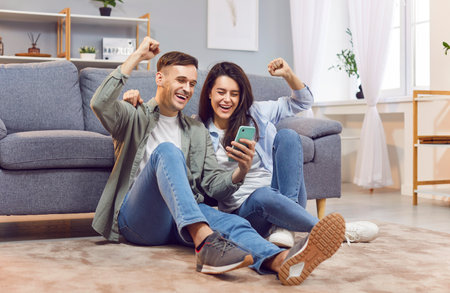 Young excited overjoyed couple sitting on floor at home holding smart phone celebrating online good news, lottery win, victory moment, invitation, internet sale, getting prize, discount, messageの写真素材