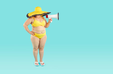 Funny woman in yellow bikini, beach flip flops, big wide brimmed sun hat standing on blue empty blank copy space background, holding megaphone, making announcementの写真素材