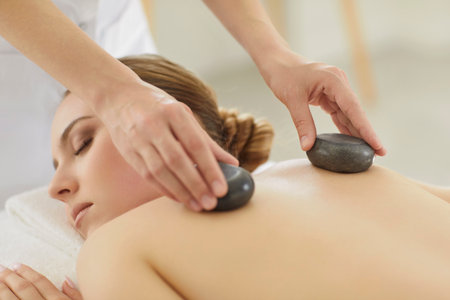 Portrait of pretty young woman with closed eyes lying relaxing in spa salon getting massage therapy with hot stones. Therapist doing back treatment procedure. Wellness and beauty day conceptの写真素材