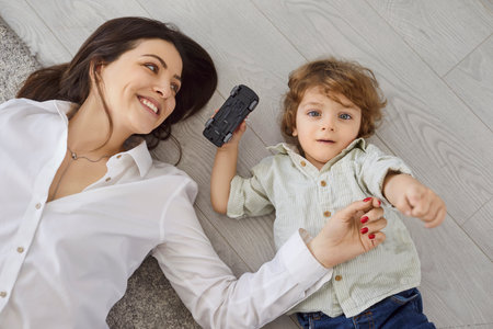 Happy mother and son lying on floor and playing together, top view. Family fun time of parent and kid at home, young mom smiling, child holding toy car to play. Bonding and love, imagination conceptの写真素材