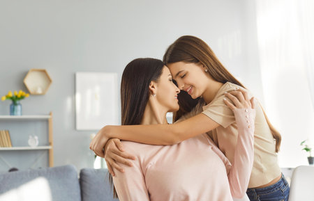 Loving teenage daughter gently hugging her mother sitting on sofa. Happy girl leaning forehead to smiling woman, sharing warm affection and deep emotional connection, showing care and family bonds.の写真素材