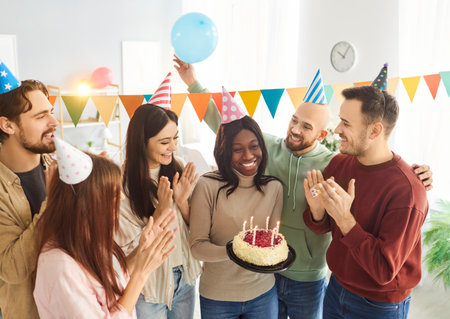 Portrait of a smiling young happy african american woman holding her birthday cake with candles standing with cheerful friends clapping indoors. Birthday party and celebration concept.の写真素材