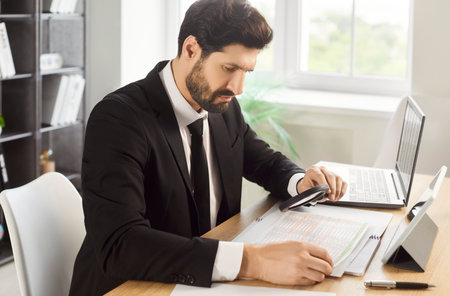Analysis of business documents by busy businessman with magnifying glass in office. Accountant holding magnifier to look at paper report, investigate and control financial risks. Audit, data conceptの写真素材