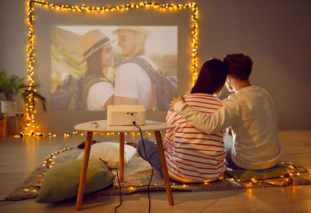 View from behind of couple watching photos with video projector. Happy family couple sitting on floor in cosy room decorated with garland lights and hugging in front of projector wallの写真素材