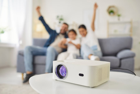 Happy family watching movie with projector at home. Digital device with lens on round table, excited father, mother and child sitting on sofa in living room to watch TV show or film togetherの写真素材
