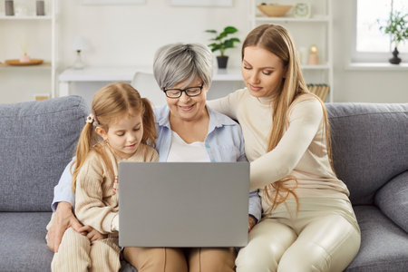Three female generations family fun at home online with laptop computer, cute little granddaughter girl, mother, grandmother, beloved granny, funny grandchild and young mom enjoy leisure time togetherの写真素材
