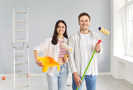 Young couple happy painters planning new flat construction, painting wall with paint roller brush, wearing protective uniform finishing task for room moving, building repair, remodeling and renovationの写真素材