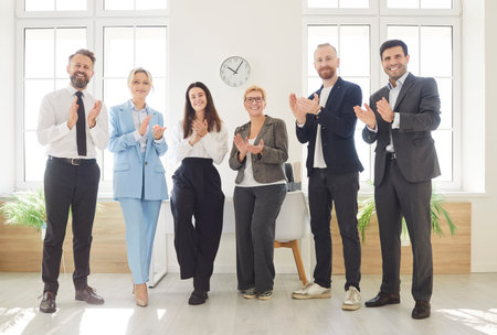 Group of business professionals standing in bright office space, smiling and clapping their hands. Full length portrait of happy colleagues showing support and appreciation, celebrating work success.の写真素材