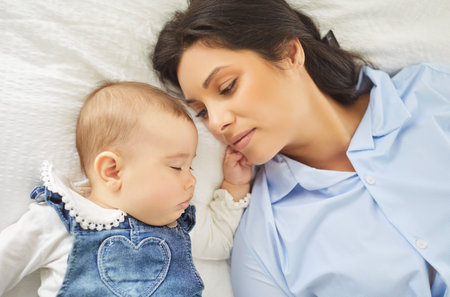 Close portrait of a mother gazing at her sleeping infant on a white bed, showing maternal love and...