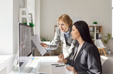 Portrait of young confident business women employees sitting at the desk on workplace, looking at pc computer monitor screen discussing company finance data in office. Accounting job concept.の写真素材