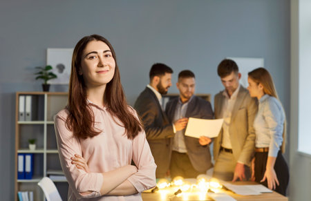 Portrait of confident young business woman looking at camera standing in office with people company...