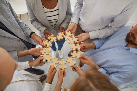 Business team joining parts of jigsaw puzzle. Group of diverse entrepreneurs or office workers standing in circle and holding wooden jigsaw pieces in hands. Teamwork concept. Top view, from aboveの写真素材
