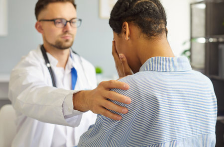 Confident young male doctor supporting female upset sick patient covering face with hands in office. Physician standing with frustrated woman during exam in medical clinic. Medicine concept.の写真素材