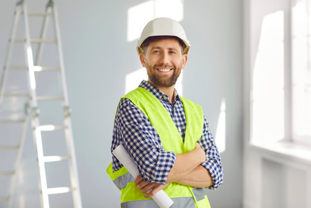 Portrait of smiling builder or architect in hardhat inside new house. Happy bearded man in hard hat and workwear standing with arms folded at home construction site with step ladder in backgroundの写真素材